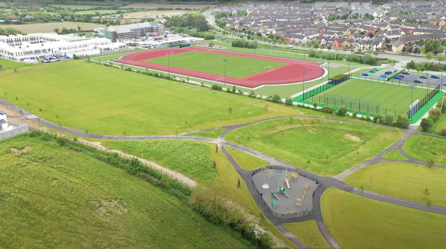 Aerial view of a sports complex with red track, green fields, and surrounding countryside and houses.