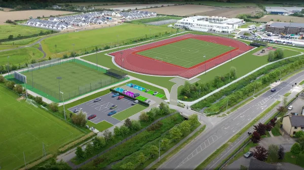 Aerial view of a sports complex with red track, green fields, parking lots, and surrounding buildings.