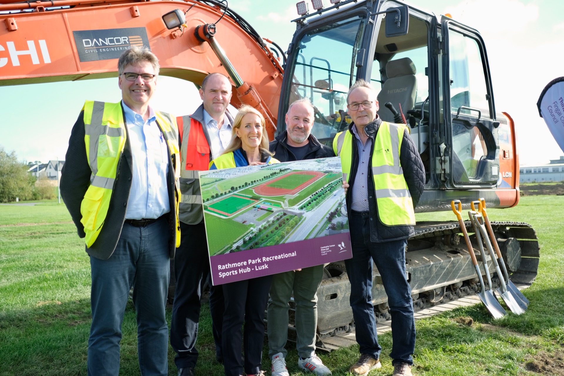 Group of workers in hi-vis vests pose beside excavator, holding site plan for Rathmore Park.