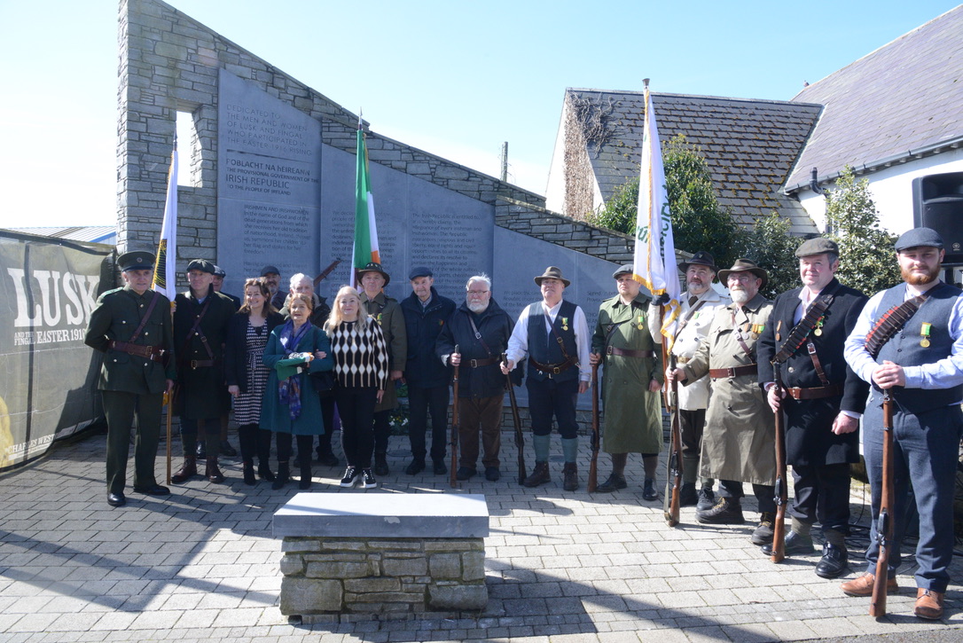 Group of uniformed veterans and civilians stand outdoors holding flags near a stone memorial wall.