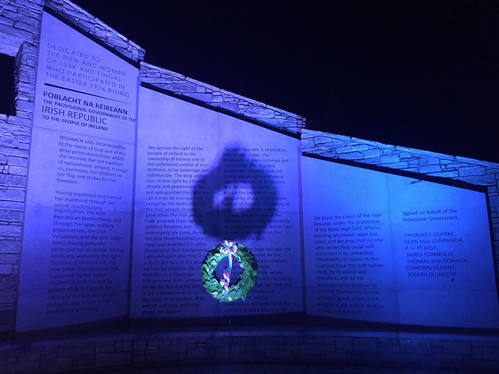 Illuminated stone memorial wall with Irish text panels and a central multicolored emblem at night.