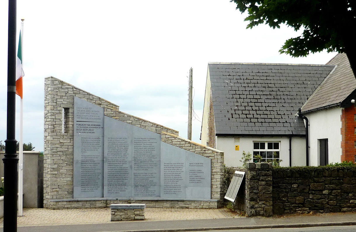 Stone memorial wall with plaques and a small bench beside a modern building under cloudy sky.