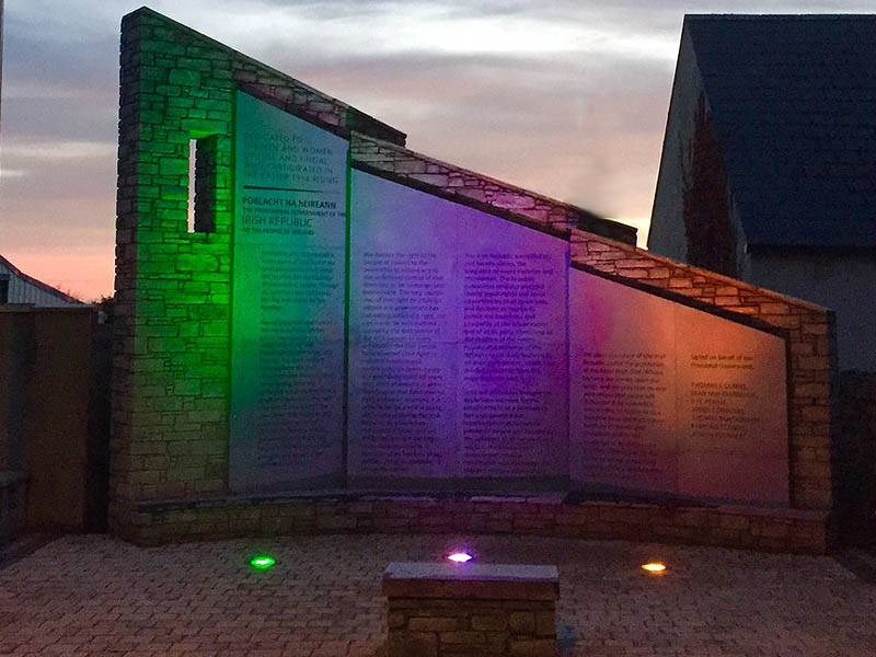 Brick memorial wall with illuminated panels in green, purple, and orange at dusk, with buildings behind.